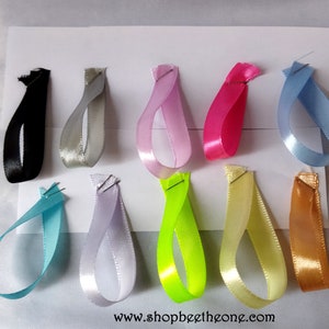 Peut inclure: Assortiment de dix bobines de ruban satiné de différentes couleurs, dont noir, argent, rose, rose vif, bleu clair, blanc, vert lime, jaune et or. Les rubans sont disposés en rangée sur une surface blanche. Le texte "www.shopbeetzone.com" est imprimé en bas de l'image.