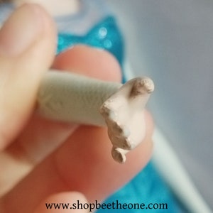 May include: A close-up of a doll's hand, painted in a light skin tone, with visible fingers and a rounded wrist. The hand is attached to a cylindrical, off-white object. The background is blurred, with hints of blue and white.