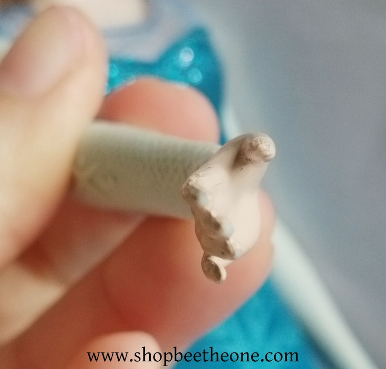 May include: A close-up of a doll's hand, painted in a light skin tone, with visible fingers and a rounded wrist. The hand is attached to a cylindrical, off-white object. The background is blurred, with hints of blue and white.
