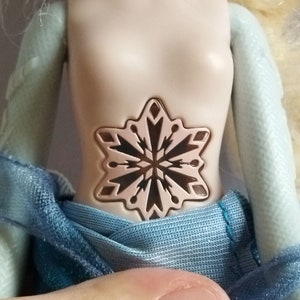 May include: A doll with a light skin tone, wearing a blue dress. The doll's torso features a snowflake design. The doll's arms are light blue. The website address www.shopbeetheone.com is visible at the bottom of the image.