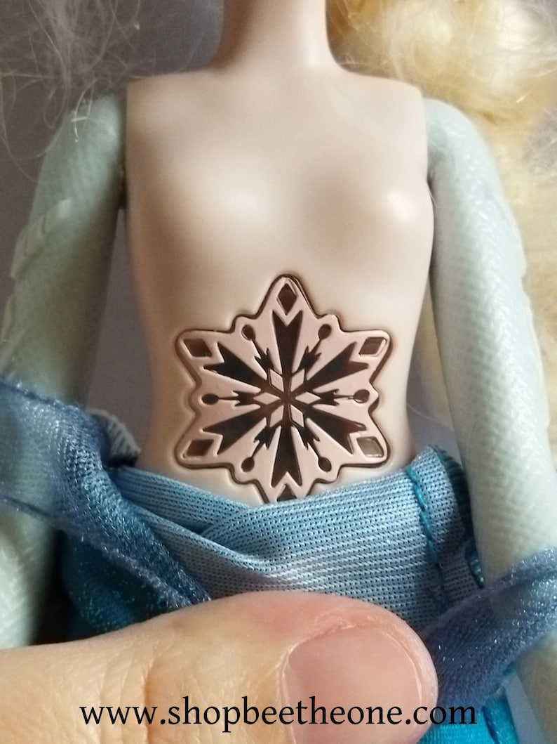 May include: A doll with a light skin tone, wearing a blue dress. The doll's torso features a snowflake design. The doll's arms are light blue. The website address www.shopbeetheone.com is visible at the bottom of the image.