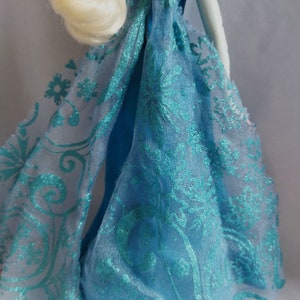 May include: A doll in a light blue dress with a sheer turquoise overlay featuring a floral and snowflake design. The doll has blonde hair in a braid. The website address www.shopbeetheone.com is visible.