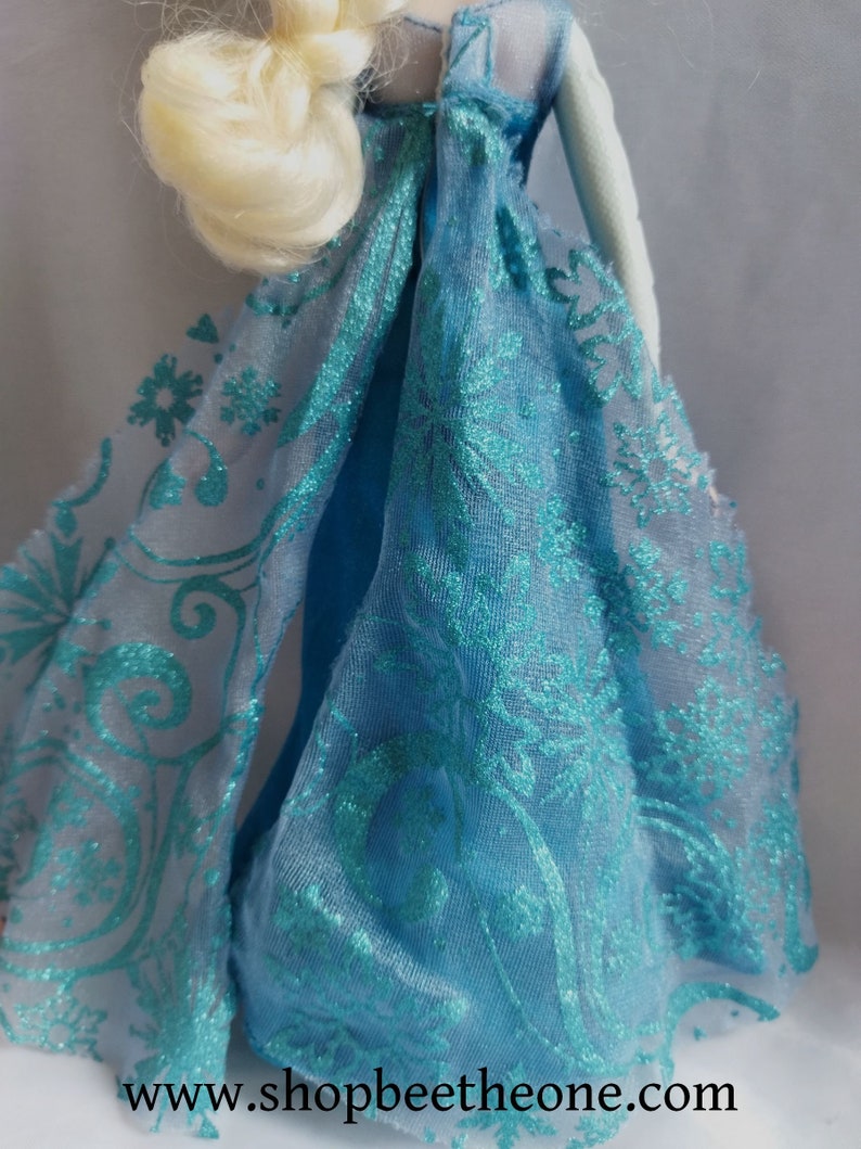 May include: A doll in a light blue dress with a sheer turquoise overlay featuring a floral and snowflake design. The doll has blonde hair in a braid. The website address www.shopbeetheone.com is visible.