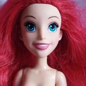 May include: A doll with red hair, blue eyes and a bright smile. The doll is wearing a nude-coloured body and has long, flowing hair.
