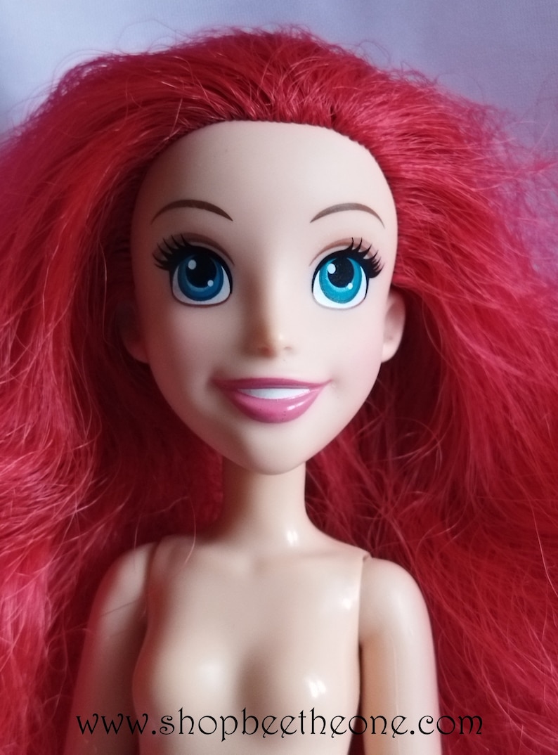 May include: A doll with red hair, blue eyes and a bright smile. The doll is wearing a nude-coloured body and has long, flowing hair.