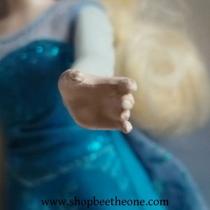 May include: A close-up of a doll's hand, with a light skin tone, reaching forward. The doll is wearing a blue dress with a sparkly bodice and a light blue sleeve. The website address www.shopbeetheone.com is visible at the bottom.
