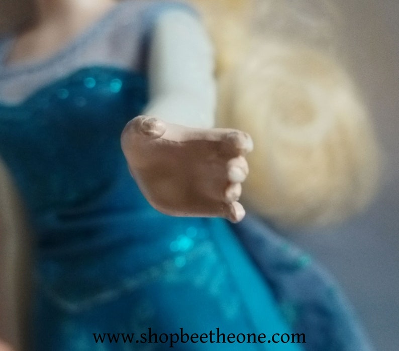 May include: A close-up of a doll's hand, with a light skin tone, reaching forward. The doll is wearing a blue dress with a sparkly bodice and a light blue sleeve. The website address www.shopbeetheone.com is visible at the bottom.
