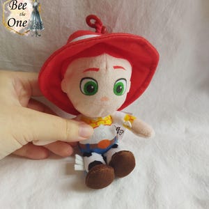 K&ouml;nnte beinhalten: Ein Pl&uuml;schtier des Charakters Jessie aus der Toy Story-Reihe. Jessie tr&auml;gt ihren charakteristischen roten Cowboyhut, ein wei&szlig;es Hemd mit einem gelben Sheriffstern, einen blauen Jeansrock mit einem wei&szlig;en Kuhmuster und braune Stiefel. Sie hat rote Haare, gr&uuml;ne Augen und einen freundlichen Gesichtsausdruck.