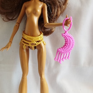 May include: A doll with brown skin and long brown hair is holding a pink plastic crescent moon shaped accessory. The doll is wearing a gold belt with a decorative design.