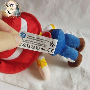 K&ouml;nnte beinhalten: Ein roter und wei&szlig;er Pl&uuml;sch-Cowboyhut mit einer blauen und braunen Pl&uuml;sch-Cowboyfigur. Das Spielzeug hat ein Etikett mit der Aufschrift "Whitehouse Leisure International Ltd Basildon, Essex, SS14 3BX, UK غسيل يدوي فقط يجفف طبيعيا . صنع في الصين I Wash Only. Dry Naturally. Made in China 12m+ CE".
