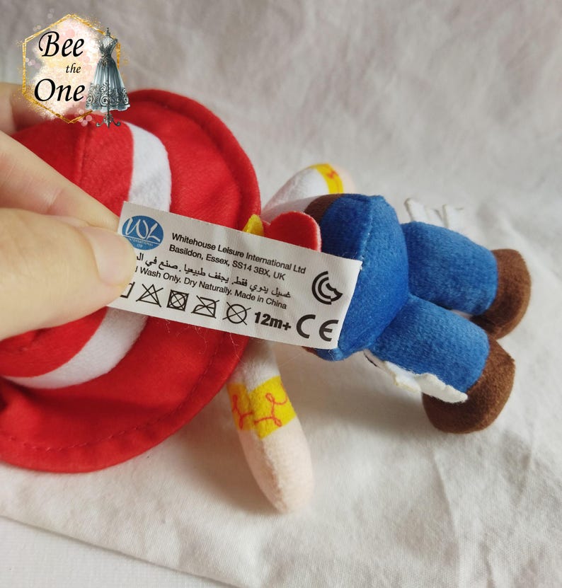 K&ouml;nnte beinhalten: Ein roter und wei&szlig;er Pl&uuml;sch-Cowboyhut mit einer blauen und braunen Pl&uuml;sch-Cowboyfigur. Das Spielzeug hat ein Etikett mit der Aufschrift "Whitehouse Leisure International Ltd Basildon, Essex, SS14 3BX, UK غسيل يدوي فقط يجفف طبيعيا . صنع في الصين I Wash Only. Dry Naturally. Made in China 12m+ CE".