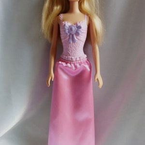 May include: A blonde doll wearing a pink dress with a purple flower detail on the bodice. The doll is wearing pink shoes.