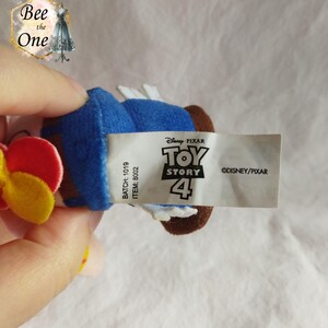 K&ouml;nnte beinhalten: Ein blaues Pl&uuml;schtier von Woody aus Toy Story 4. Das Spielzeug hat eine rote und gelbe Blume auf dem Kopf. Das Etikett auf dem Spielzeug lautet "Disney Pixar Toy Story 4" und "&copy;DISNEY/PIXAR".