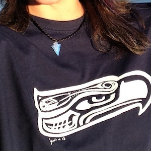 May include: A navy blue t-shirt with a white graphic of a stylized eagle head. The eagle's eye is visible and the graphic is outlined in black. The shirt has a black cord necklace with a blue arrowhead pendant.