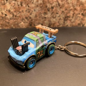 May include: A blue toy race car keyring with the number 72 on the side. The car has a spoiler that says "Surge Fly" and a silver key ring. The car has blue and green accents.