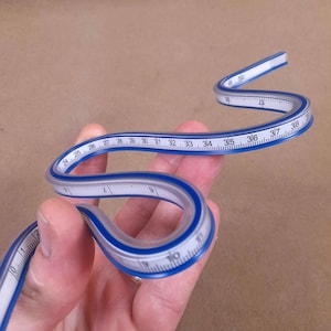 Può includere: Un righello curvo flessibile e pieghevole con superficie bianca, segni di misurazione neri e bordi blu. Il righello è piegato a forma di S e mostra incrementi numerici da 0 a 50, offrendo uno strumento versatile per il disegno tecnico.