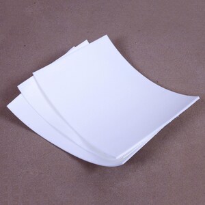 May include: A stack of three white, curved paper sheets. The sheets have a textured surface and are arranged in a fan-like pattern, with the top sheet slightly overlapping the others. The background is a solid, neutral brown.