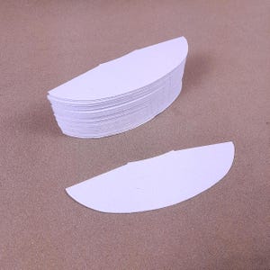 May include: A stack of white, semi-circular paper cutouts, with one cutout lying separately. The cutouts are arranged on a brown surface. The paper appears to be thick and textured.