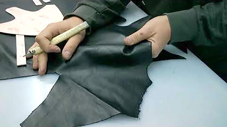 Puede incluir: Una persona corta cuero negro con un cuchillo artesanal. El cuero se sujeta en su lugar mientras se usa el cuchillo para cortar alrededor de un patr&oacute;n de papel. La escena sugiere artesan&iacute;a en cuero o un oficio similar.