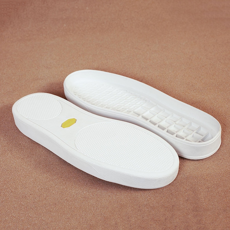 May include: Two white shoe insoles with textured surfaces and a yellow oval detail. One insole is upside down, revealing the patterned sole. The insoles are designed for sneakers or casual shoes.