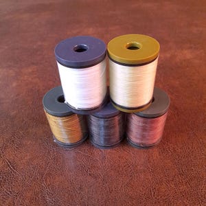 May include: A stack of six spools of thread in various colors, including white, gold, and dark grey. Each spool has a dark-colored top with a central hole. The thread is tightly wound around the spool.