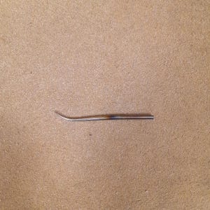 May include: A slender, metallic tool with a curved, pointed tip and a straight, cylindrical shaft. The metal exhibits a subtle heat-treated discoloration, transitioning from a silver tone at the tip to a darkened, brownish hue along the body.