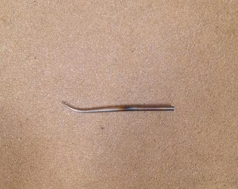 May include: A slender, metallic tool with a curved, pointed tip and a straight, cylindrical shaft. The metal exhibits a subtle heat-treated discoloration, transitioning from a silver tone at the tip to a darkened, brownish hue along the body.