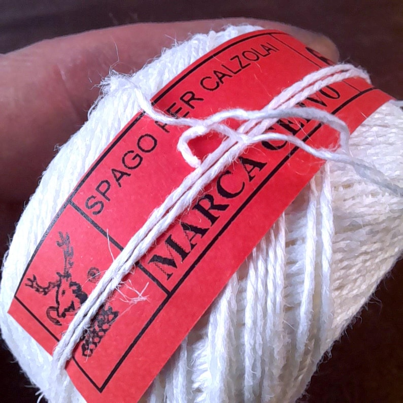May include: A ball of white yarn with a red label wrapped around it. The label has the text "SPAGO PER CALZOLA" and "MARCA CERVO" printed on it in black. A white string is tied around the yarn and label.