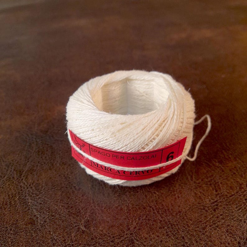 May include: A ball of off-white twine with a red label wrapped around it. The label has white text that reads "SPAGO PER CALZOLAI" and "MARCA CERVO" with the number "6". The twine is likely used for crafting or shoemaking.