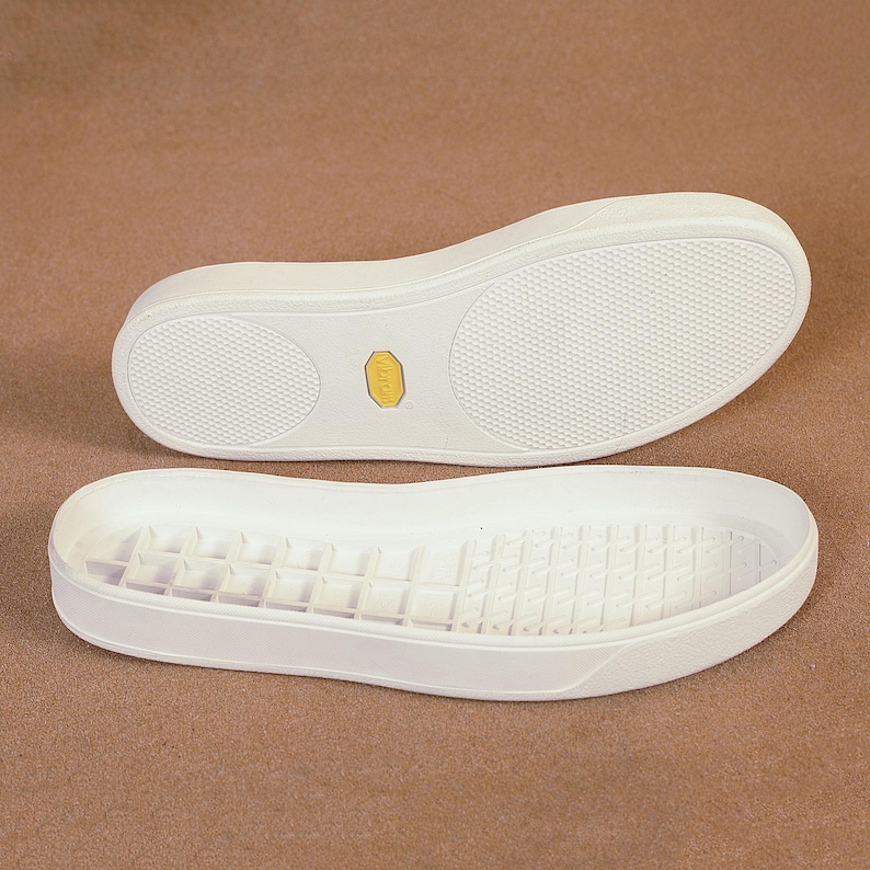 May include: Two white sneaker soles with textured bottoms are displayed against a neutral background. The soles have a rounded toe and a low profile. A small yellow logo is visible on the sole of one shoe.