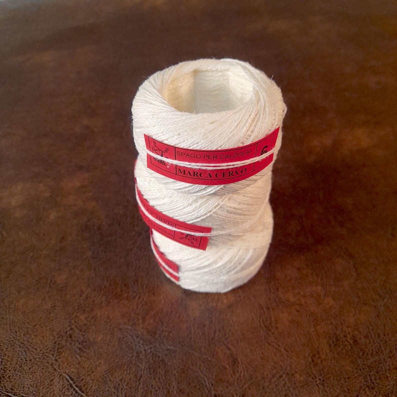 May include: Three stacked spools of white yarn with red labels. The labels read "SPAGO PER CALZOLA" and "MARCA CERVO". The yarn is tightly wound, and the spools are resting on a brown surface. The image is well-lit, highlighting the texture of the yarn.