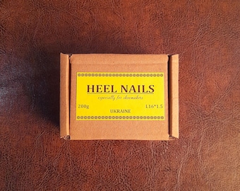 May include: A small, rectangular cardboard box with a yellow label that reads "HEEL NAILS" and "especially for shoemakers." The label also indicates a weight of 200g and the country of origin, Ukraine. The box is on a brown leather-like surface.