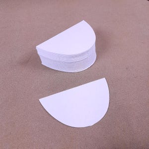 May include: A stack of white, semi-circular paper filters, with one filter separated from the stack. The filters are on a brown surface. The filters are likely for coffee or tea.