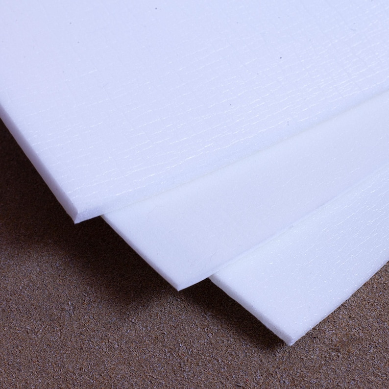 May include: Close-up of several white foam sheets, stacked at an angle. The sheets have a slightly textured surface and are likely used for packaging or crafting. The edges of the foam are visible, showing the thickness of the material.