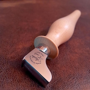 May include: A close-up of a leather working tool with a wooden handle. The tool is made of metal and has a curved blade. The handle is made of light wood and has a metal band around it. The tool is engraved with the words "Sharpo Tools" and "Made in Ukraine since 2019."