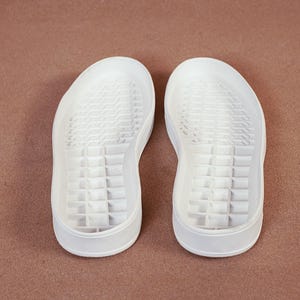 May include: Two white sneaker soles are displayed against a brown background. The soles are made of a solid material with a textured pattern on the bottom. The soles have a rounded toe and a slightly raised heel.