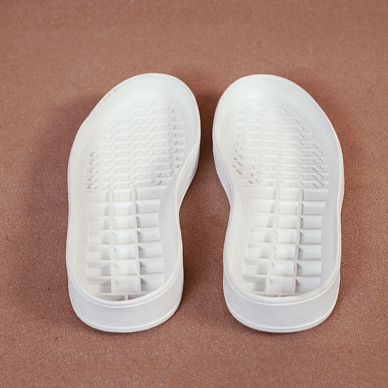 May include: Two white sneaker soles are displayed against a brown background. The soles are made of a solid material with a textured pattern on the bottom. The soles have a rounded toe and a slightly raised heel.