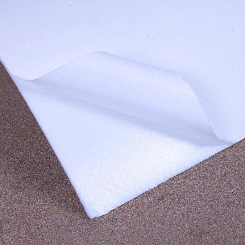 May include: A close-up of a white, textured sheet of paper with a corner folded over, revealing a smooth, bright surface. The paper rests on a brown surface, creating a contrast in textures and colors.