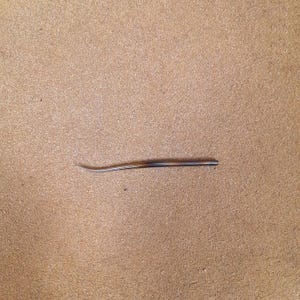 May include: A single, slender, curved needle with a pointed tip, featuring a smooth, tapered body with a gradient of brown and tan tones. The needle is positioned horizontally, showcasing its elongated, slightly arched shape and polished finish.