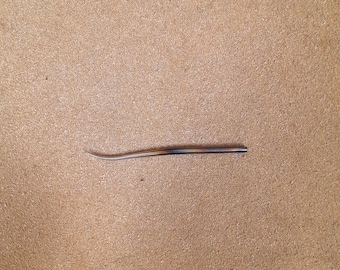 May include: A single, slender, curved needle with a pointed tip, featuring a smooth, tapered body with a gradient of brown and tan tones. The needle is positioned horizontally, showcasing its elongated, slightly arched shape and polished finish.