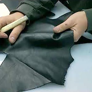 May include: A person is shown cutting black leather with a craft knife. The leather is being held in place while the knife is used to cut around a paper pattern. The scene suggests leathercraft or a similar craft.