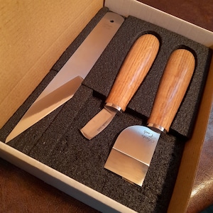 May include: A set of three stainless steel woodworking tools with wooden handles, including a long blade, a curved blade, and a square blade. The tools are in a cardboard box with black foam inserts. The text "Harke Tools" is visible on the blades.