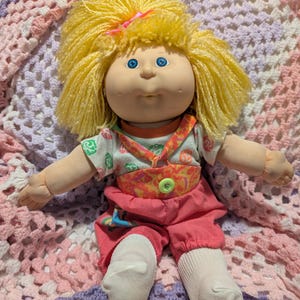 May include: A doll with bright yellow yarn hair tied with a pink bow, blue eyes, and dimpled cheeks. The doll wears a white short-sleeved shirt with a colourful pattern, a coral-coloured overall-style outfit with a green button, and white socks.