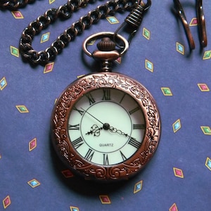 May include: A vintage-style pocket watch with a copper-toned chain and a white face with black Roman numeral markings. The watch face reads "QUARTZ".