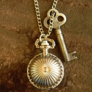 May include: A silver necklace with a silver key charm and a silver pocket watch charm. The pocket watch charm has a sunburst design.