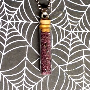 Pode incluir: Um colar de corrente prateada com um pequeno frasco de vidro cheio de glitter vermelho escuro. O frasco tem uma rolha de cortiça e está pendurado em uma corrente prateada.