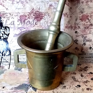 May include: Brass mortar and pestle set. The mortar is a deep bowl with a flared rim and two handles. The pestle is a smooth, round rod with a rounded end. The set is antique and has a patina finish.