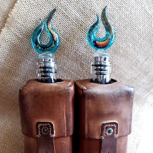 Może przedstawiać: Dwa brązowe skórzane uchwyty na butelki z niebieskimi i pomarańczowymi szklanymi korkami w kształcie płomieni.