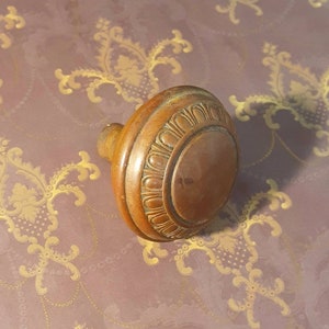 May include: A vintage, round, brass door knob with a decorative, raised pattern around the edge.