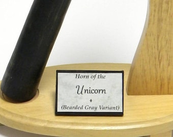 May include: A black unicorn horn on a wooden stand. The stand has a label that reads "Horn of the Unicorn (Bearded Gray Variant)".
